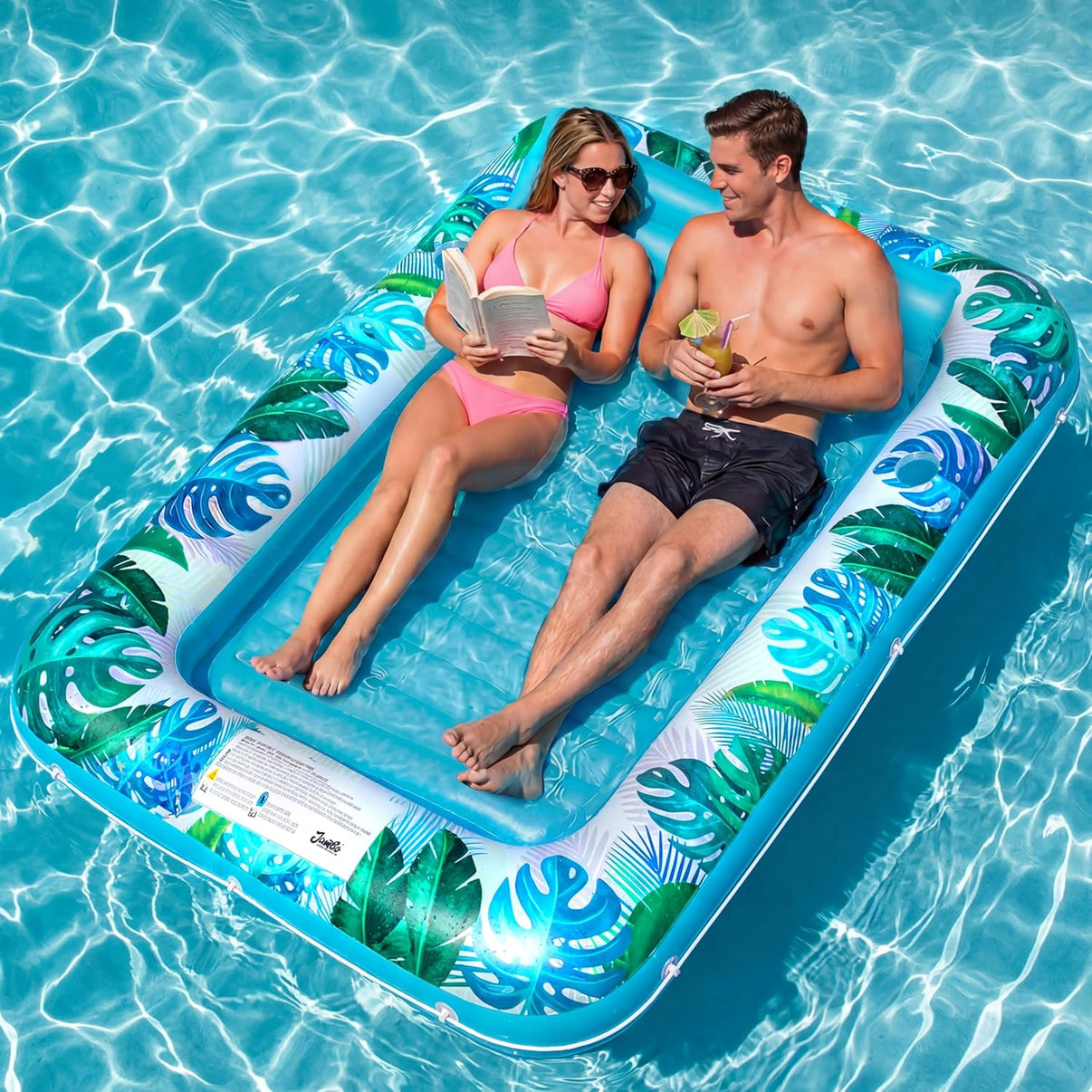Two people relaxing on a tropical-themed inflatable pool float in clear blue water.
